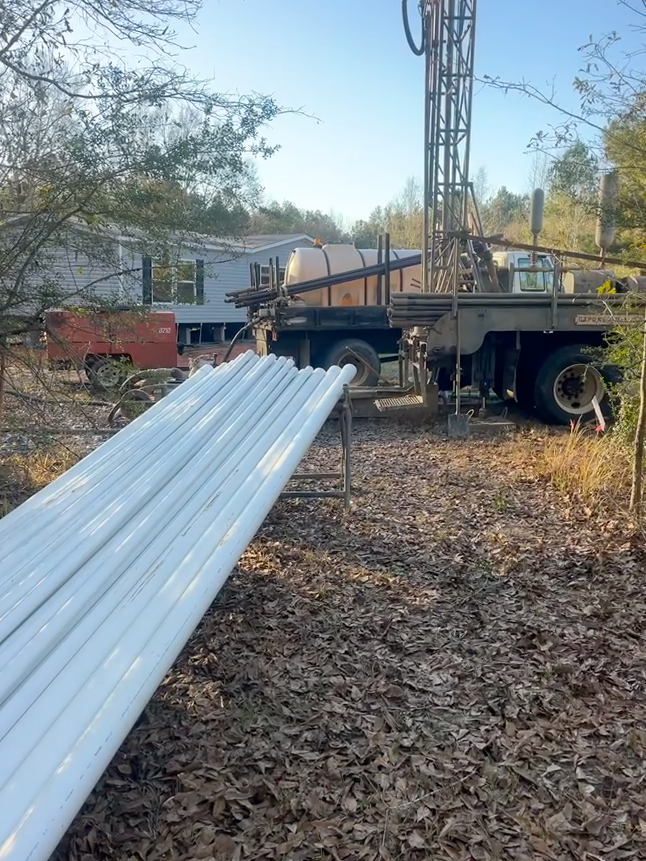 A mobile drilling rig set up on a wooded lot, with a stack of white PVC pipes in the foreground and a house nearby.