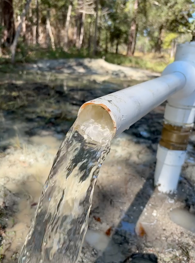 Water flows from an open white PVC pipe outdoors into a muddy, wooded area.