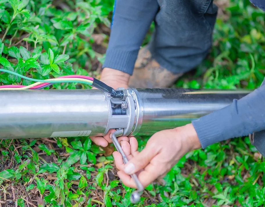 Hands tightening a metal clamp on a shiny pipe outdoors among green plants