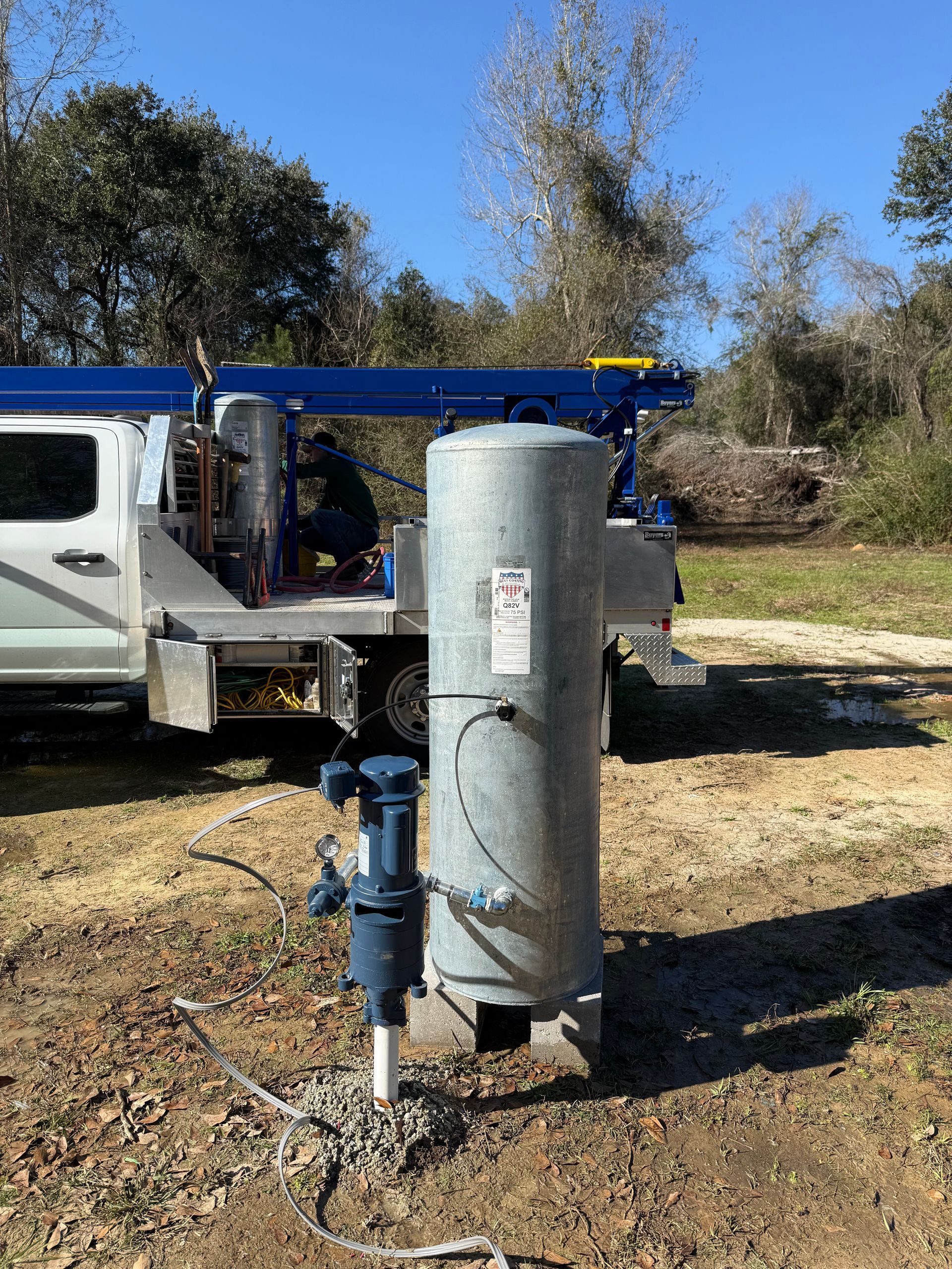 A drilling rig operates in a wooded area, discharging orange mud into a collection tank in the foreground.