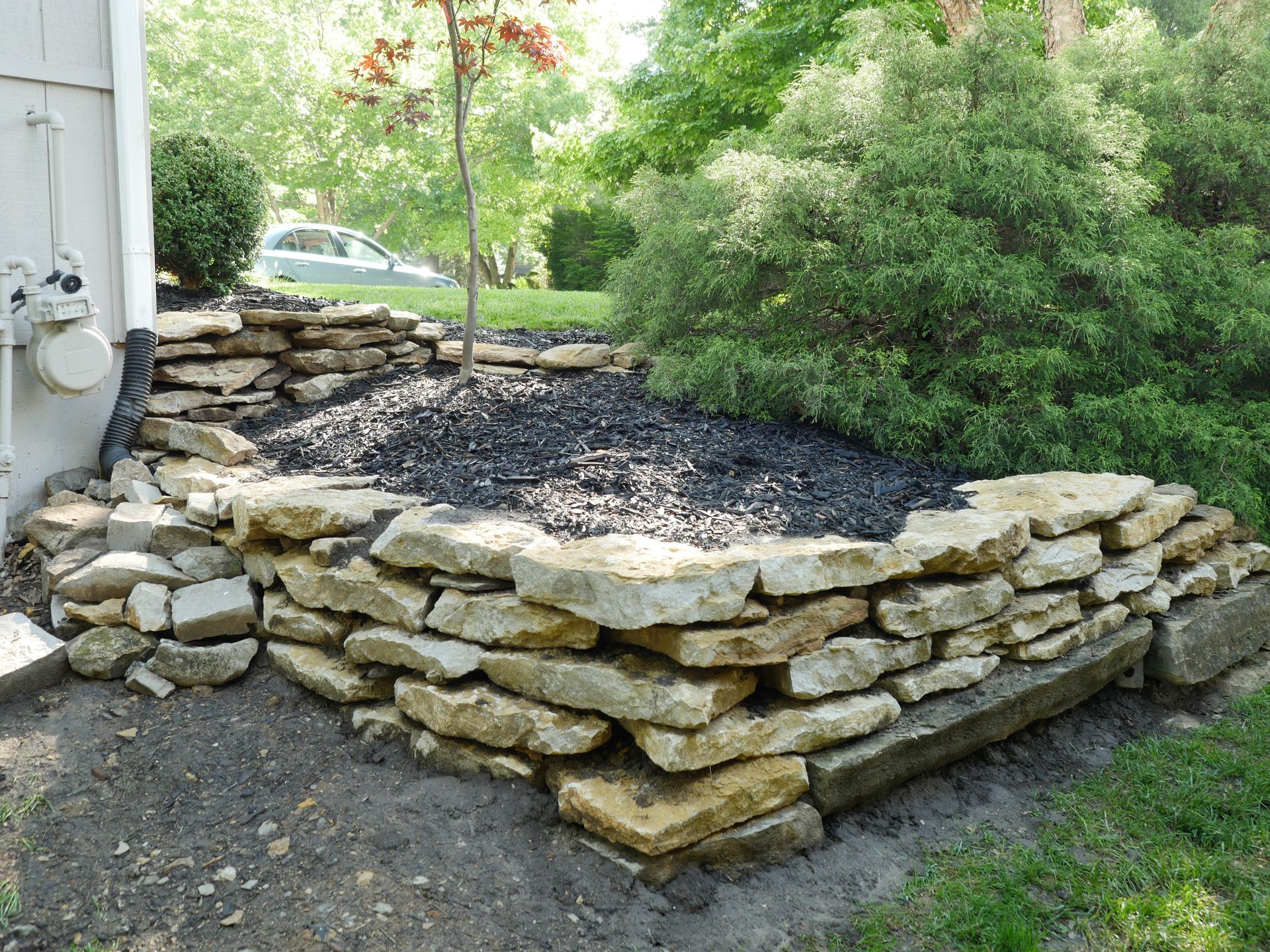A raised garden bed made of stacked tan limestone blocks filled with black mulch and a tree, located next to a house.