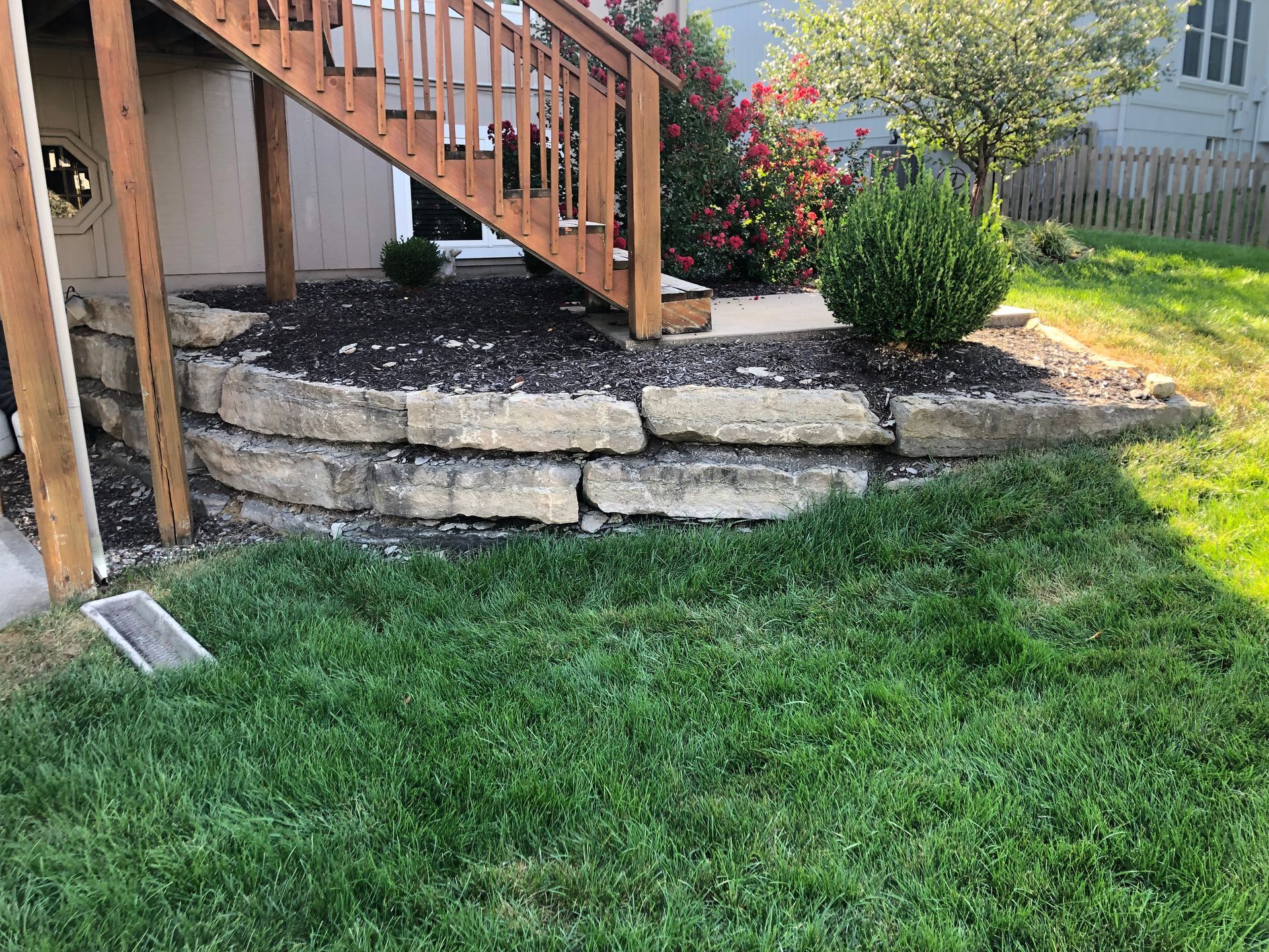 A two-tier stone retaining wall supports a garden bed beneath a wooden deck staircase in a grassy backyard.