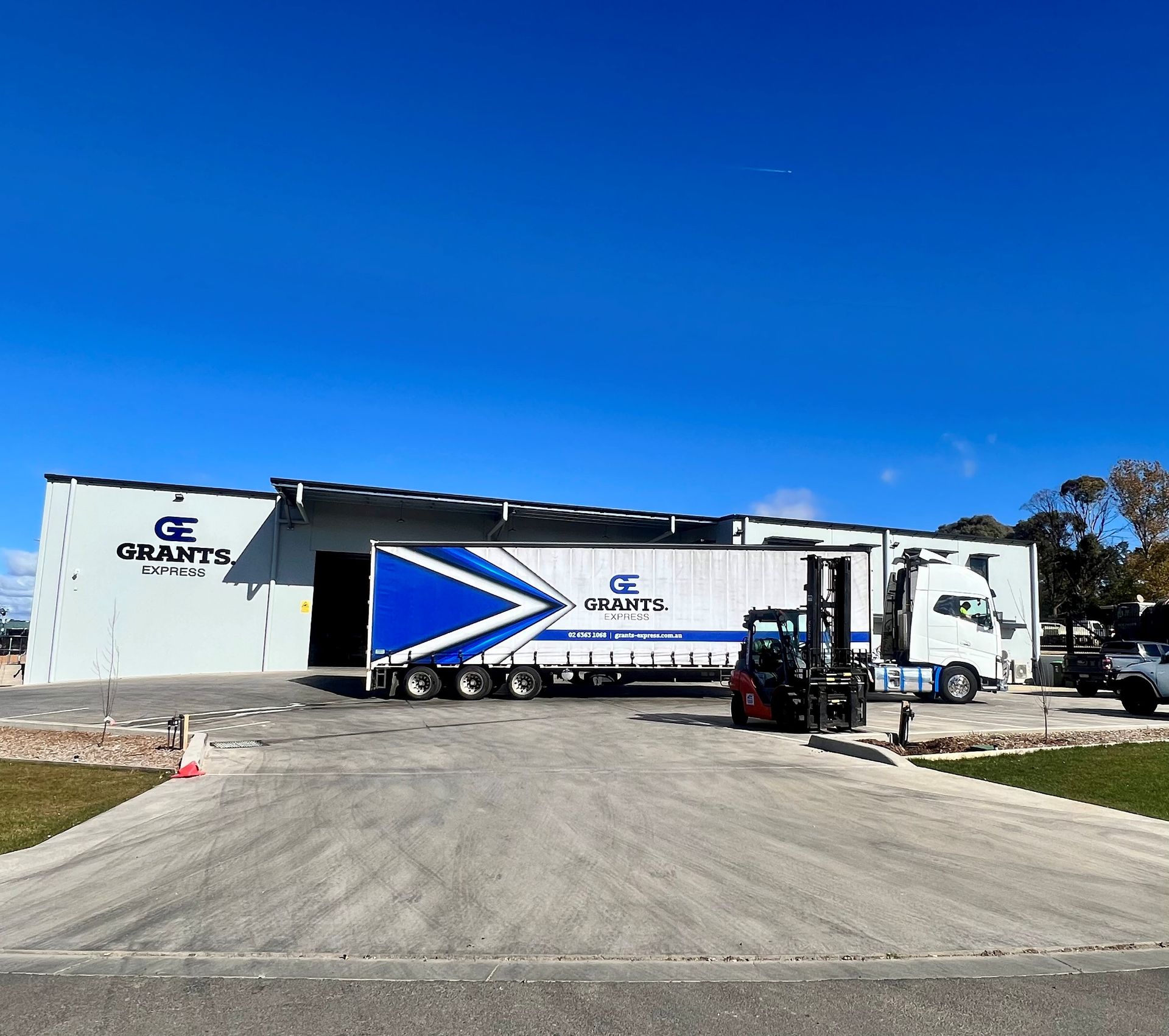 White truck trailer with blue arrow logo parked by a warehouse on a sunny day — Grants Express in Orange, NSW
