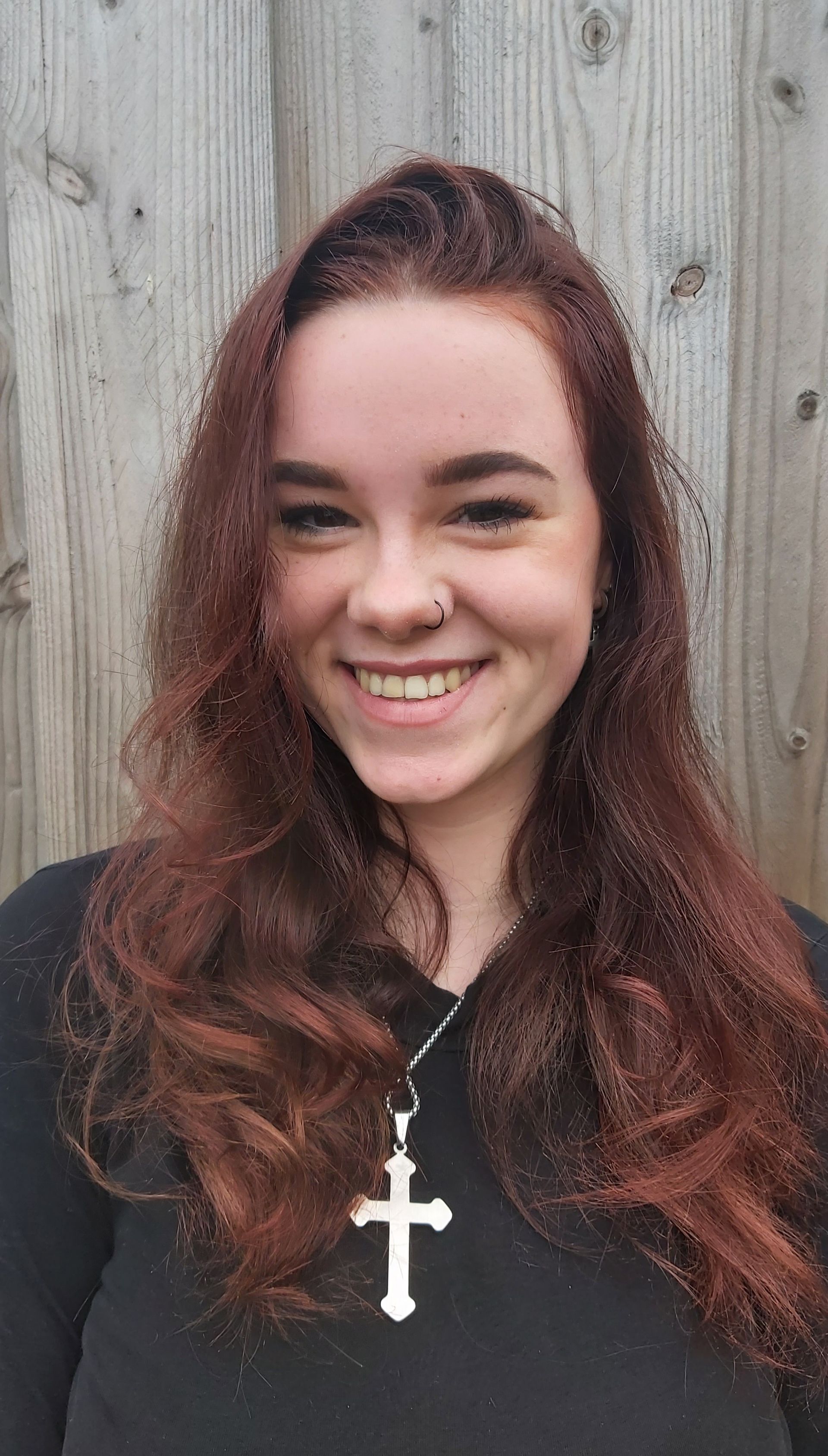 Vrouw met roodachtig haar, neusring, lachend en met een kruisje om haar nek. Voor een houten hek.