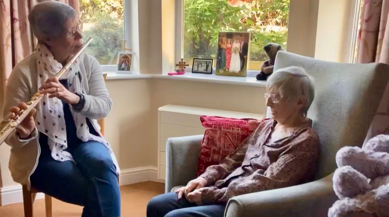 Woman plays flute for an elderly woman in a sunlit living room.