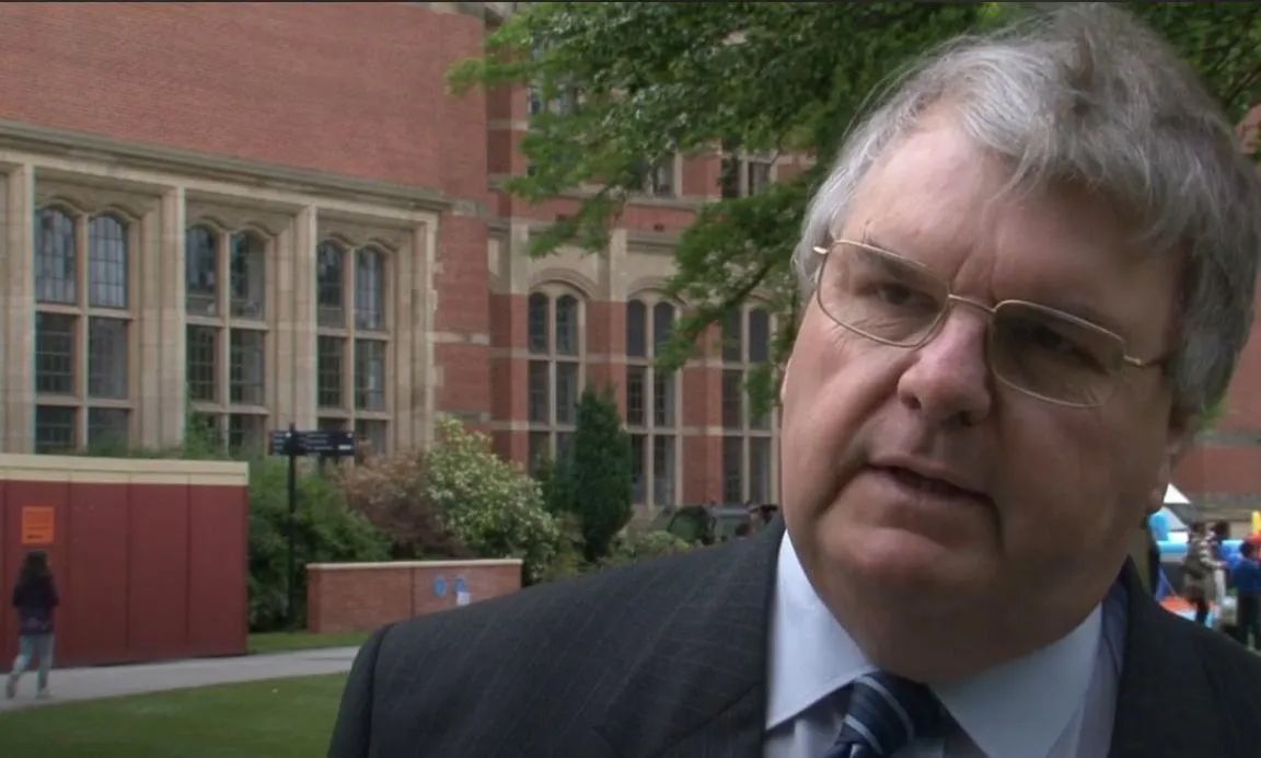Man in a suit and glasses speaking outdoors on a campus, with brick buildings in the background