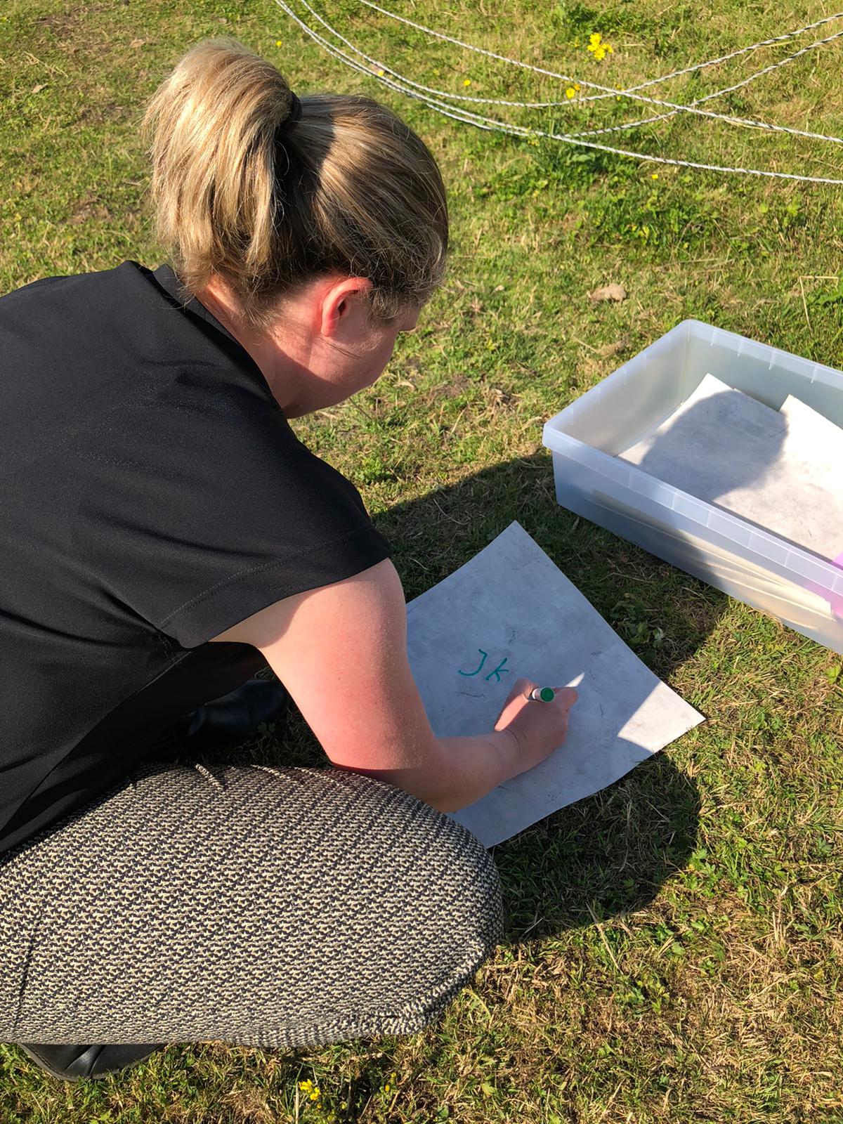 Vrouw schrijft buiten op papier; plastic afvalbak vlakbij op het gras.
