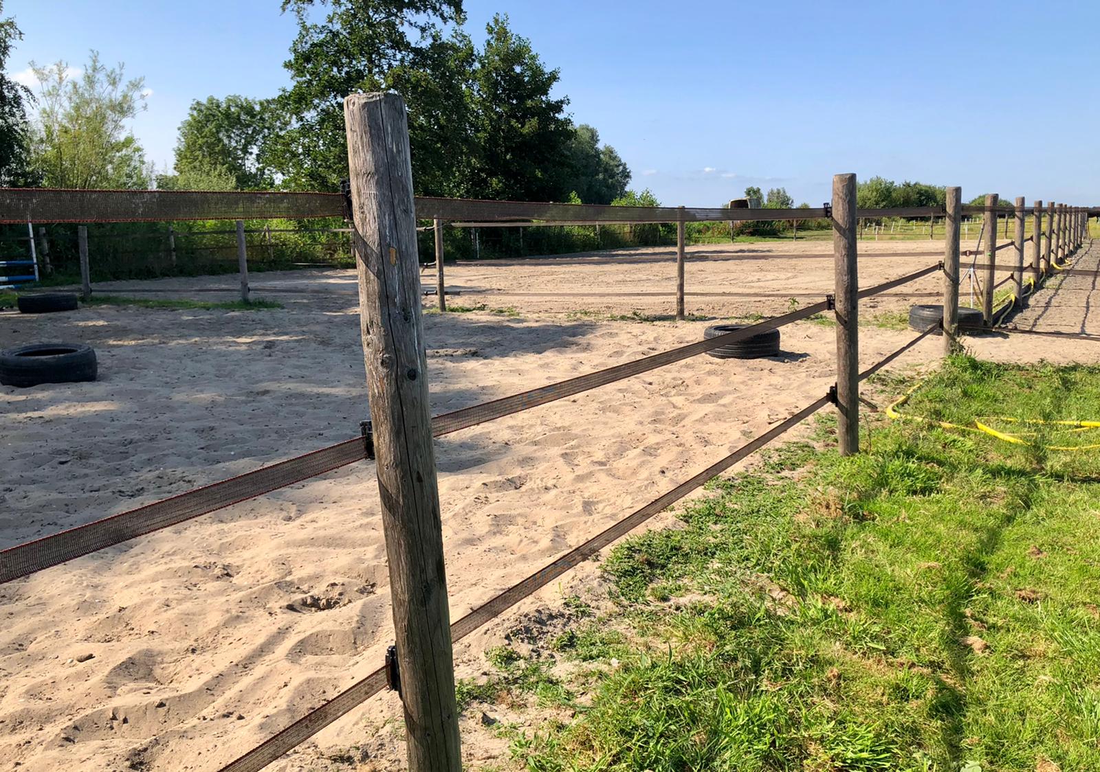 Een zanderige paardenarena met een houten hek en autobanden, op een zonnige dag.