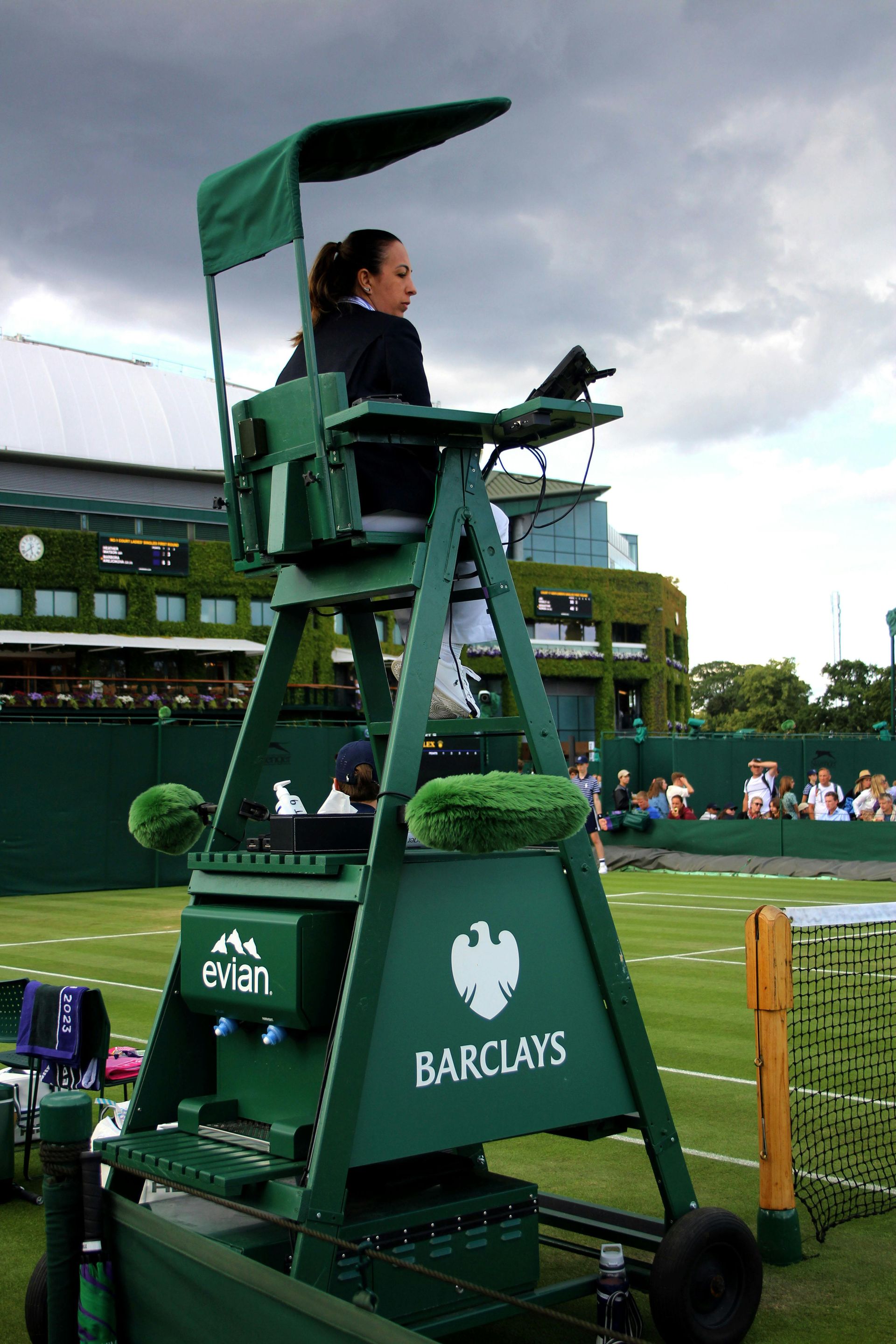 Tennis umpire in green chair at Wimbledon court, overseeing a match, under a cloudy sky.