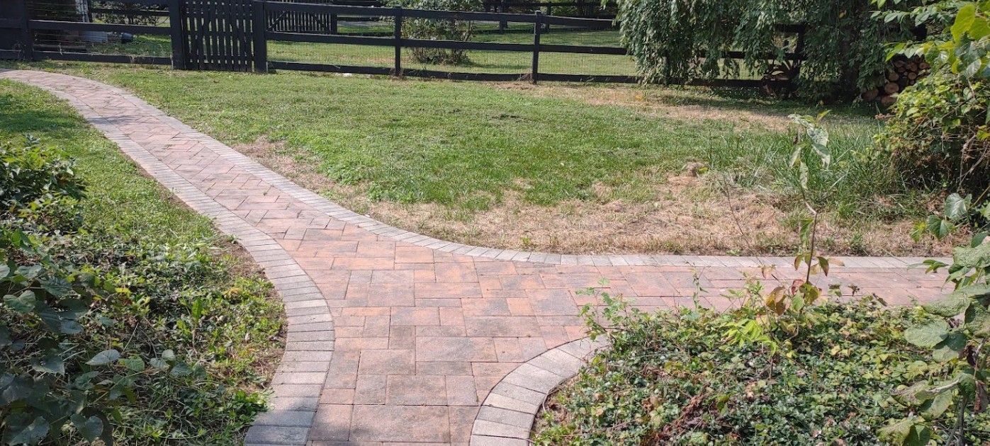A brick walkway leading to a grassy field with a fence in the background.
