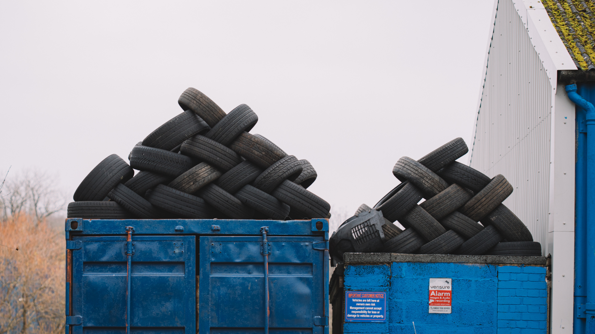 Two blue dumpsters overflowing with used tires, under a cloudy sky.