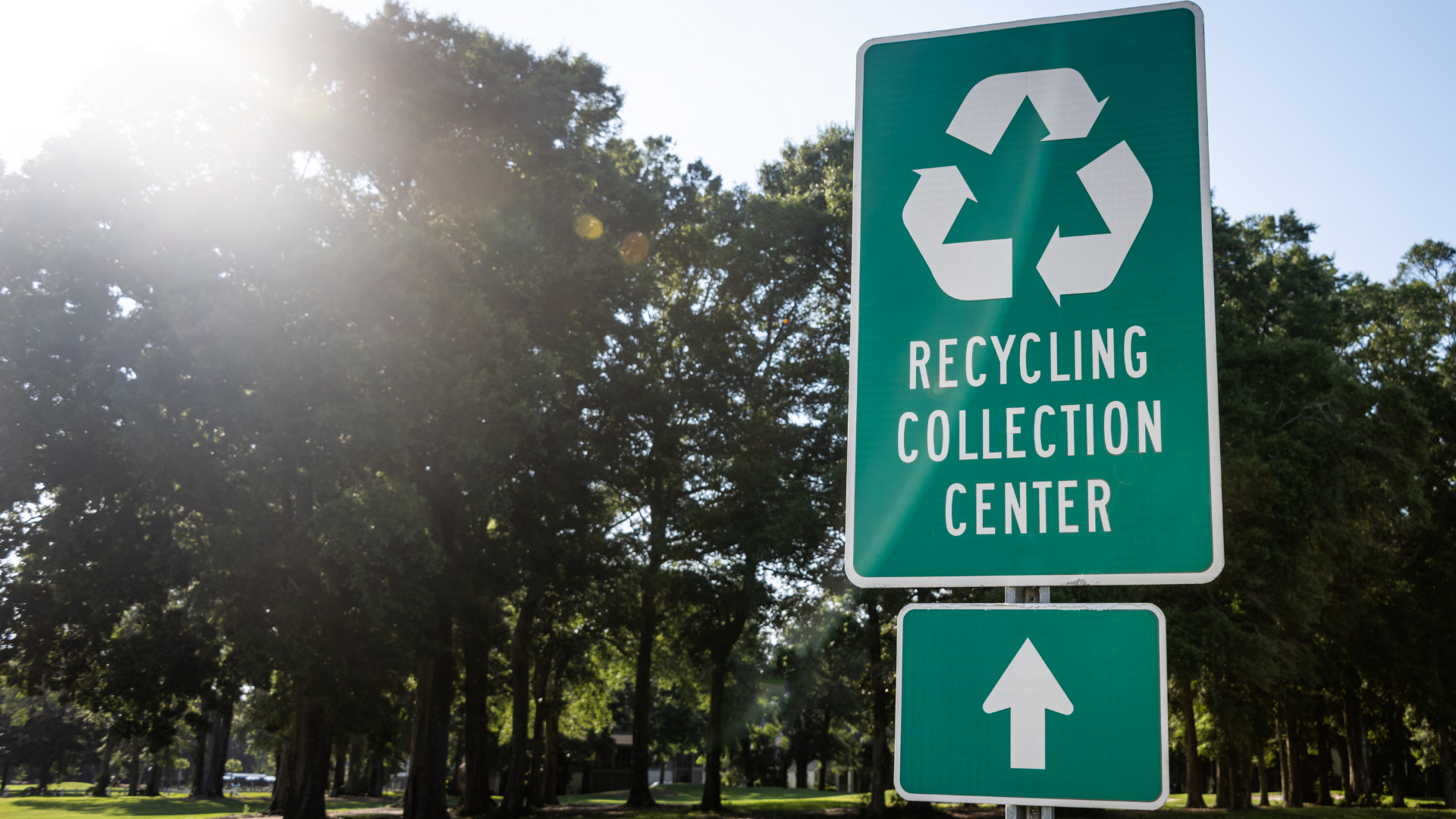 Green recycling sign with logo and arrow pointing up, in front of trees.
