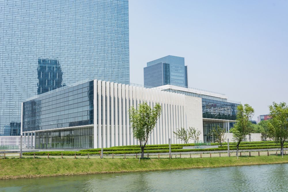 Modern glass office building beside a canal, with trees and a clear blue sky
