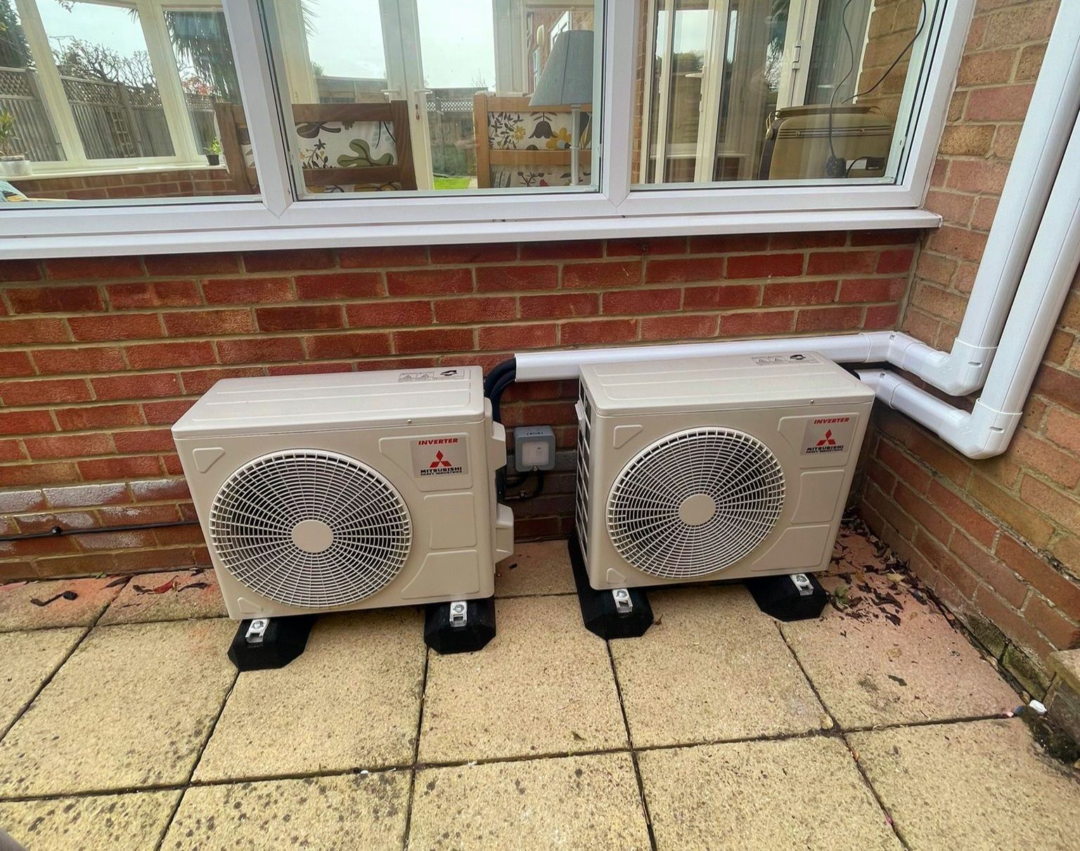 Two outdoor heat pump units beside a brick house wall and window on a paved patio.