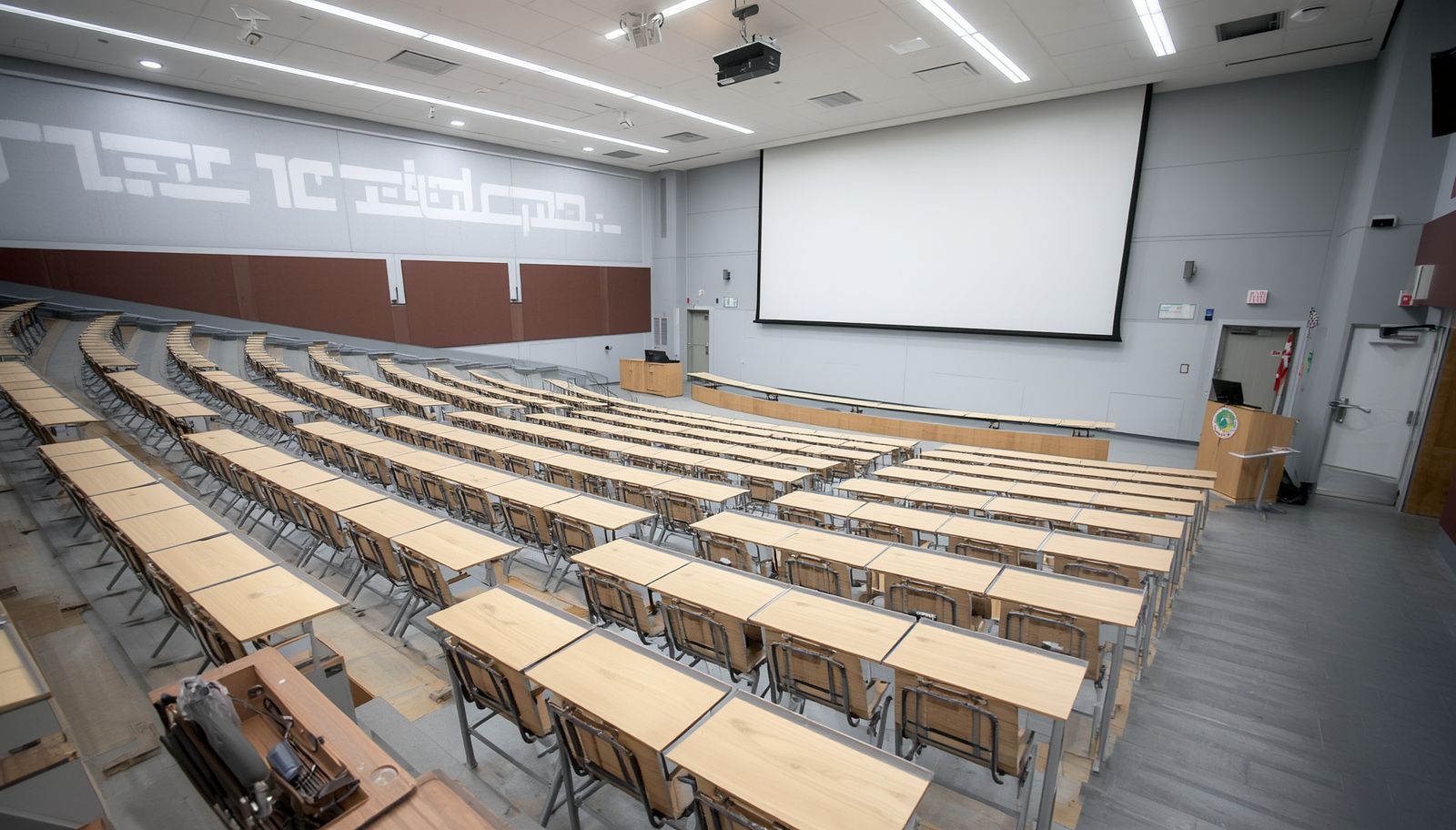 Empty lecture hall with tiered wooden desks, gray walls, and a large projector screen at the front.