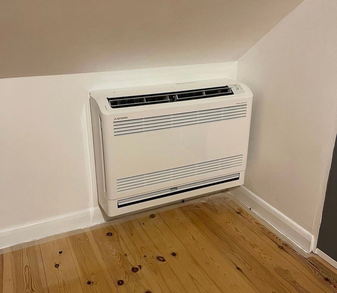 White wall-mounted heater in a corner of a room with wooden floor and sloped ceiling