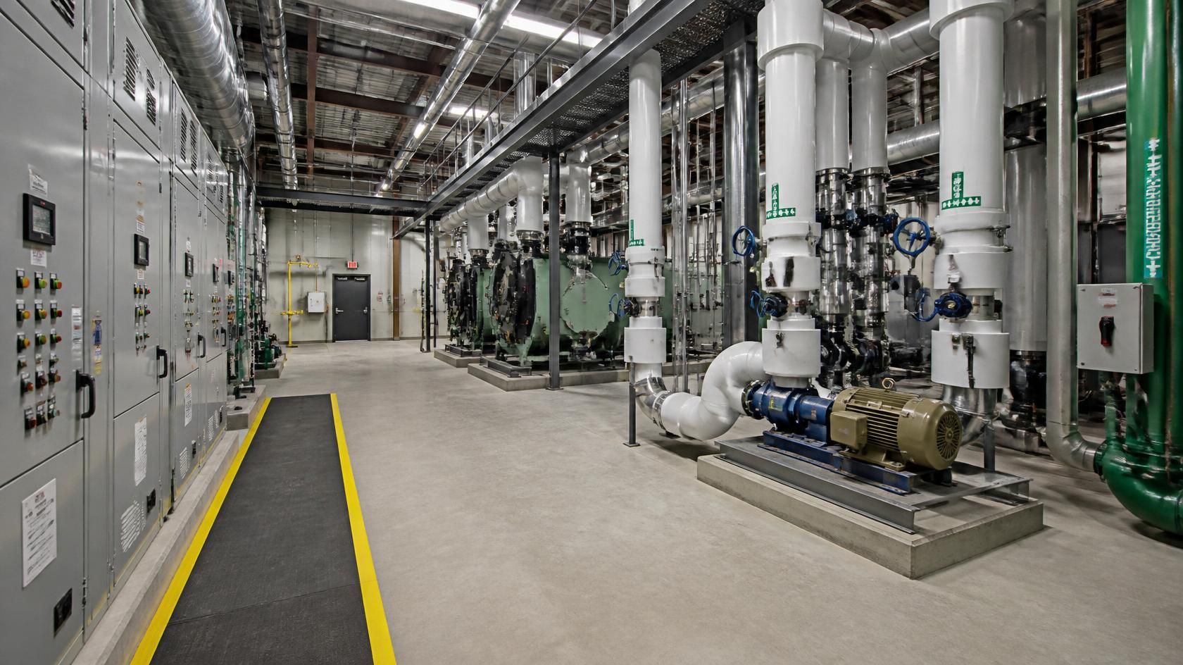 Industrial plant room with machinery, pipes, and control panels along a clean concrete aisle.