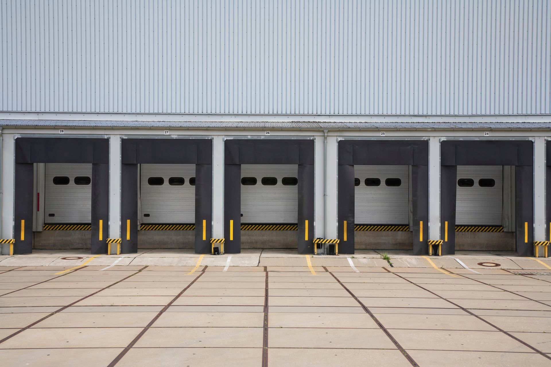 Industrial loading docks with five gray garage doors and yellow safety bollards.