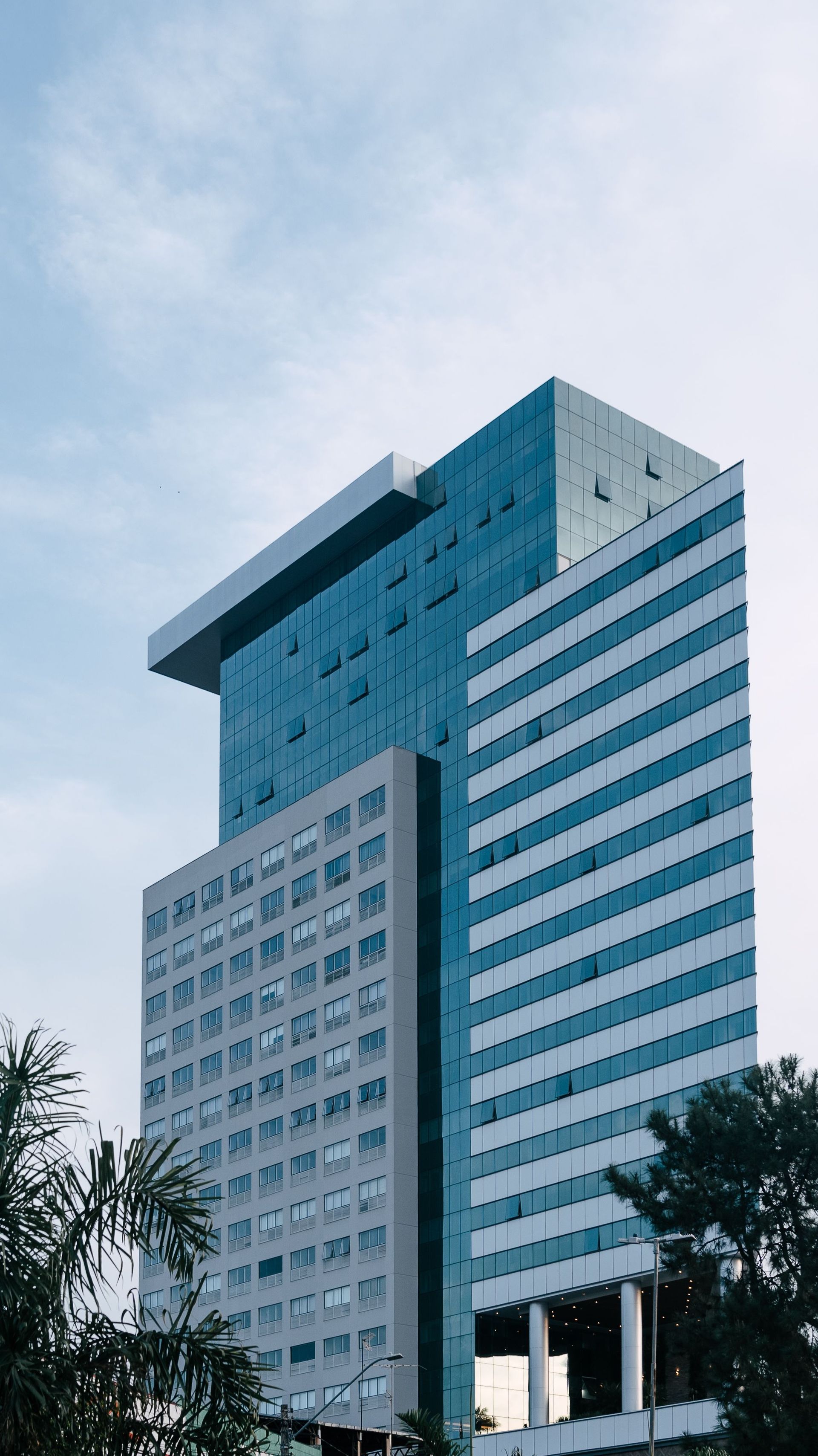 Tall modern glass office building against a cloudy sky, viewed from below with trees in the foreground.