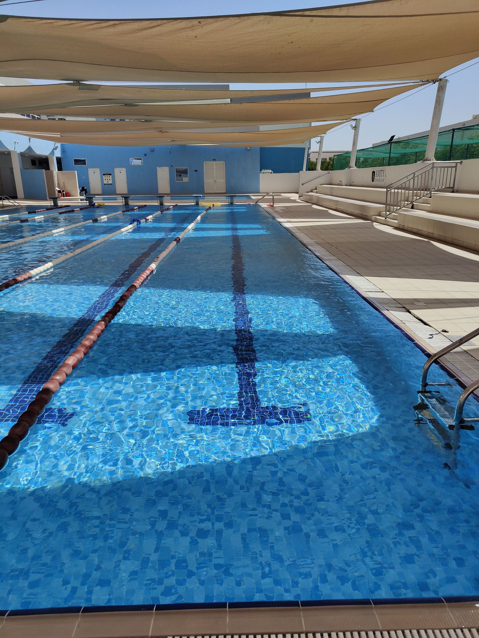 Outdoor swimming pool with lane markings, blue water, and shaded deck chairs under canopies