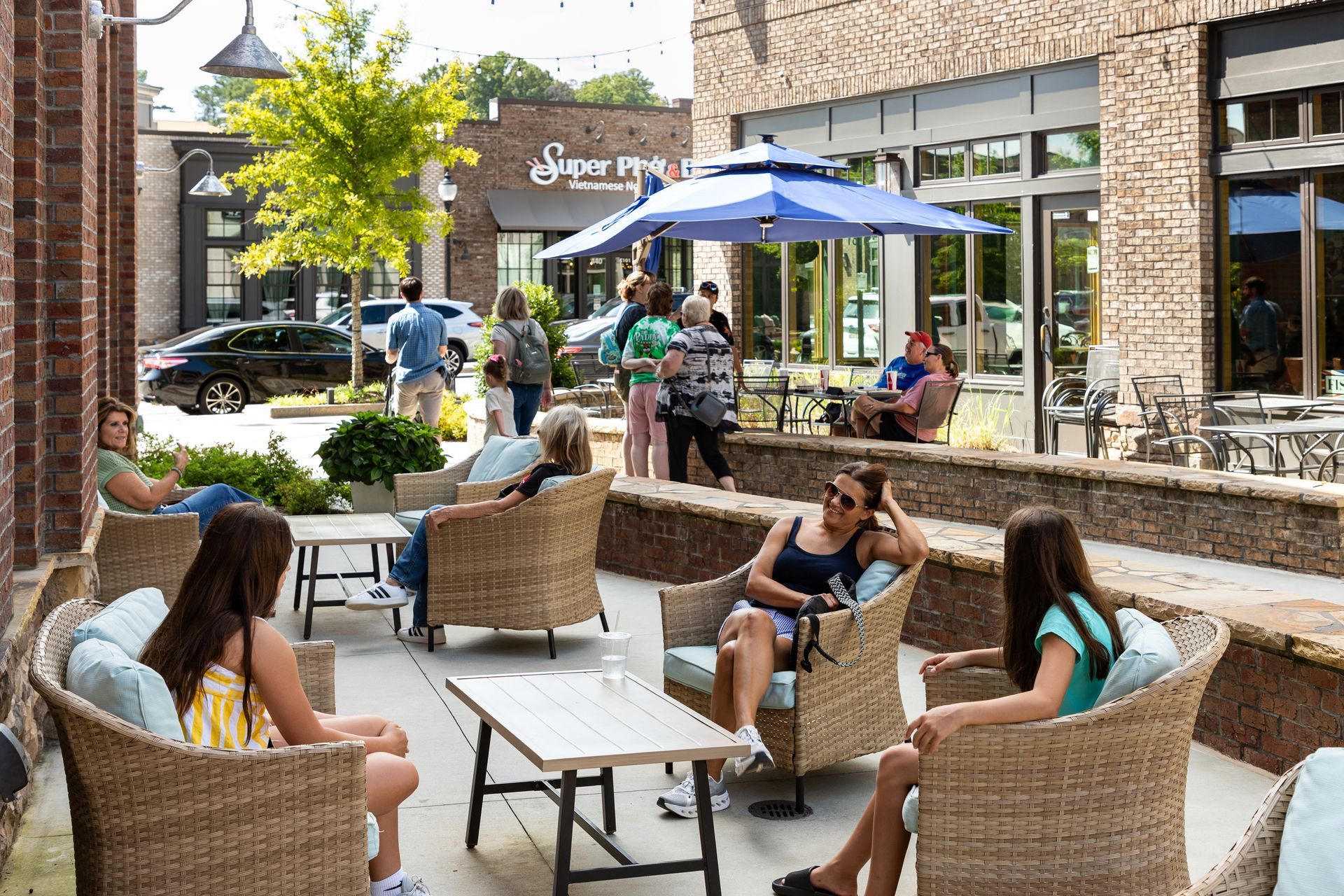 Outdoor café patio with wicker chairs, umbrellas, and people sitting and talking beside storefronts.