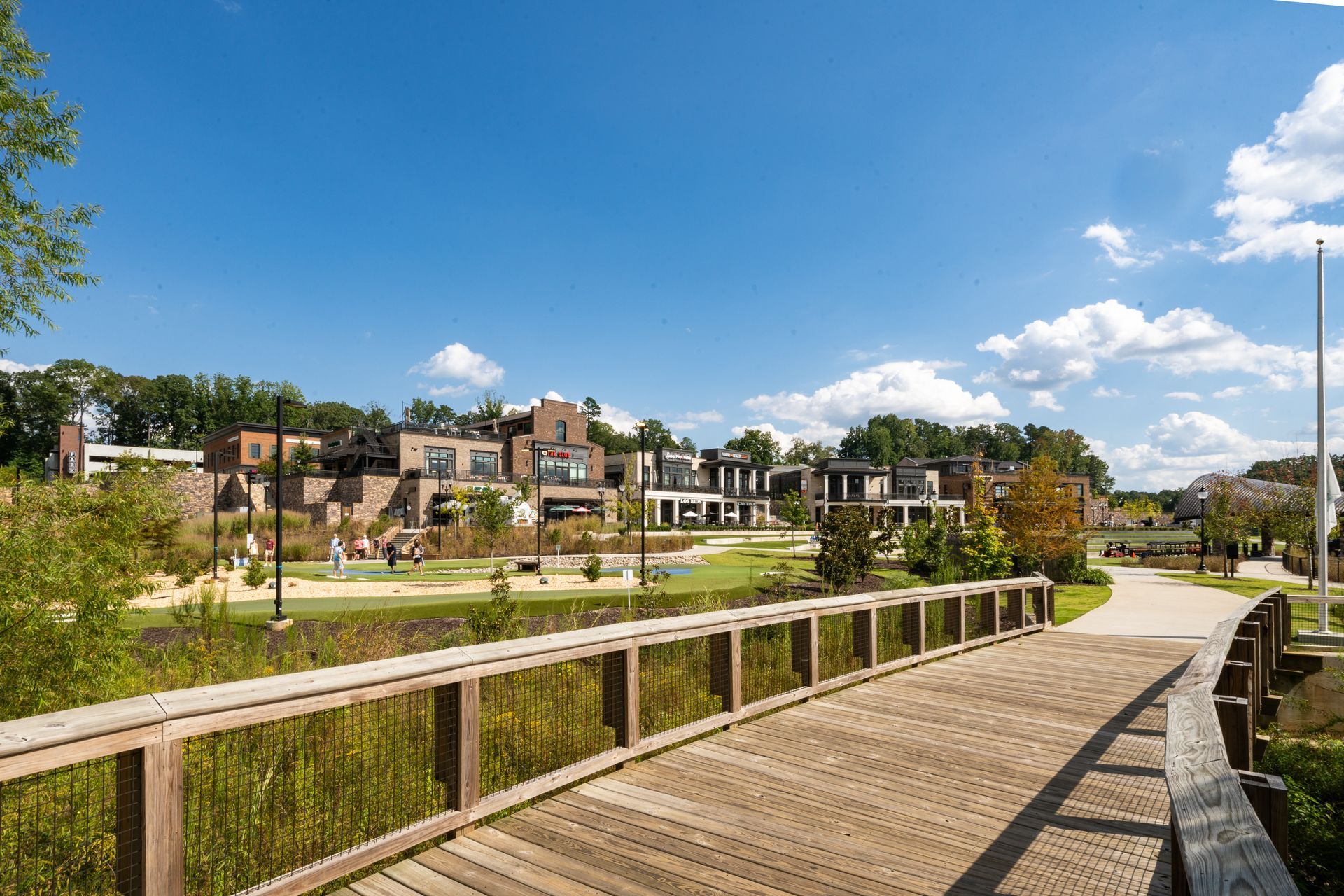 Wooden bridge over a pond in a sunny park with modern houses in the background.
