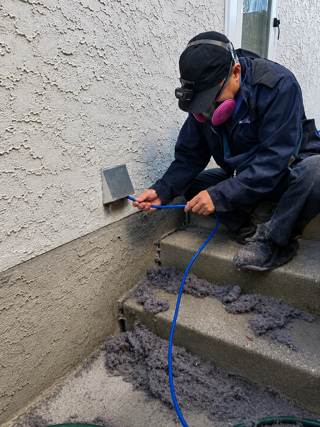 Worker cutting a small wall opening near concrete steps, using a blue hose in a narrow outdoor space