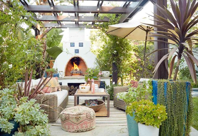 Cozy outdoor patio with an arched fireplace, lounge seating, and lush greenery under a pergola.