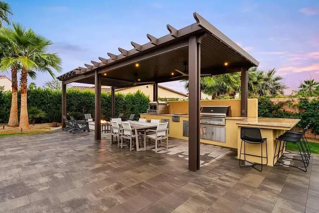 Outdoor patio with pergola, tables, chairs, and a built-in grill beside palm trees at sunset