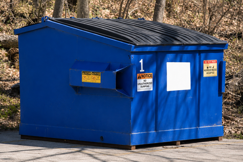 Green dumpster with black lid and warning labels on a paved area outdoors