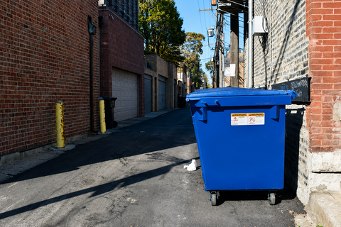 Narrow alley with brick buildings and a green dumpster in the foreground.