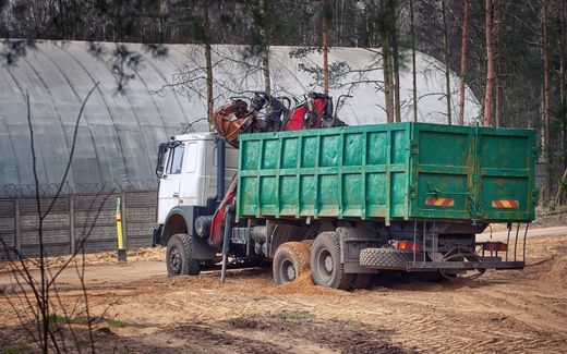 White truck with green cargo bed parked in a muddy wooded area near a greenhouse structure