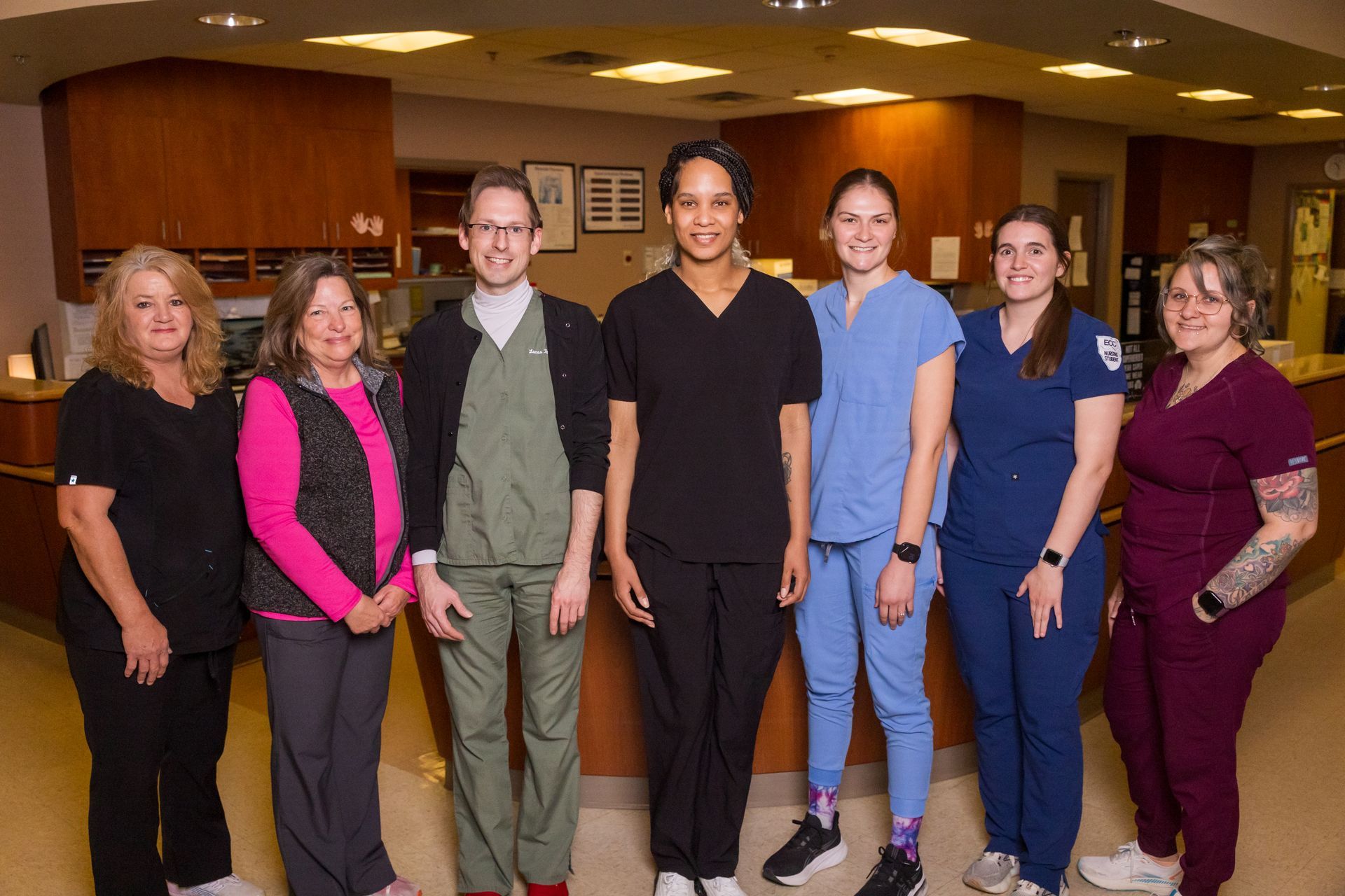 Seven healthcare workers standing in a brightly lit office, various scrubs.