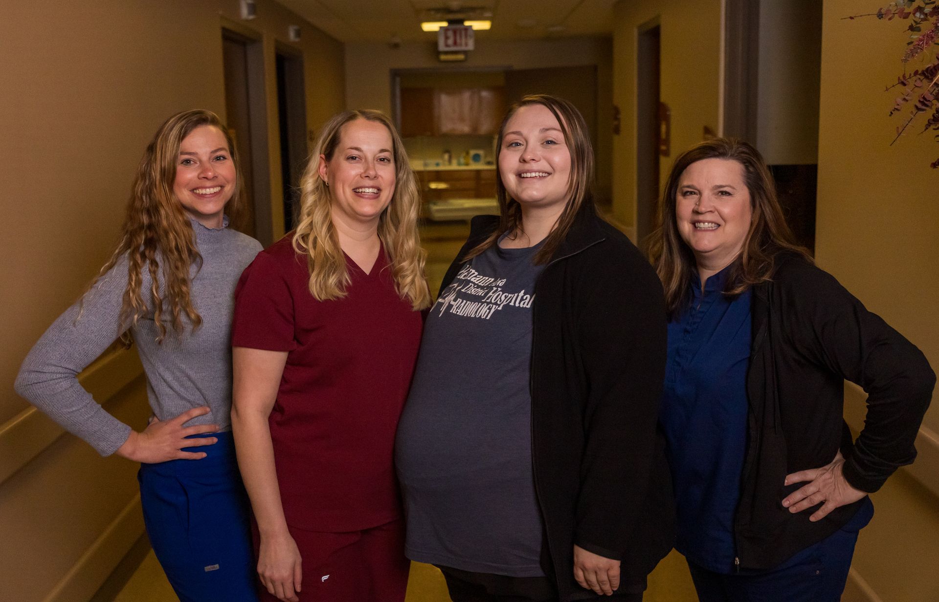 Four smiling women posing in a hallway, one with a prominent belly. They all have arms on their hips.