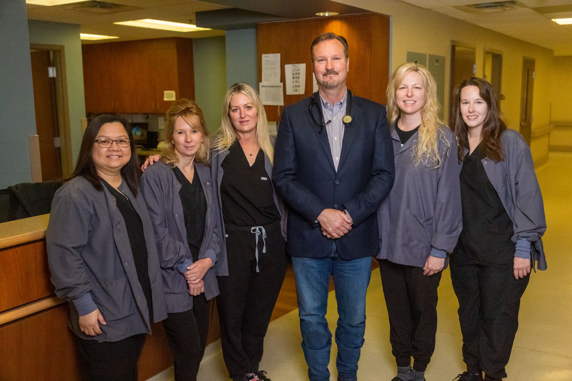 Group of six people posing in a medical facility. Five in scrubs, one in a blazer and jeans, smiling.