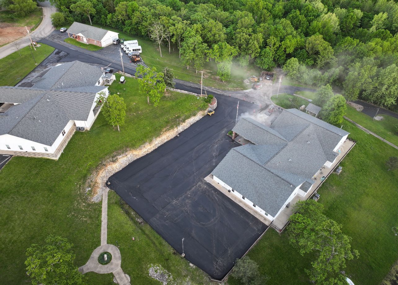 Aerial view of a suburban property with two gray buildings, blacktop parking, and trees surrounding the area.