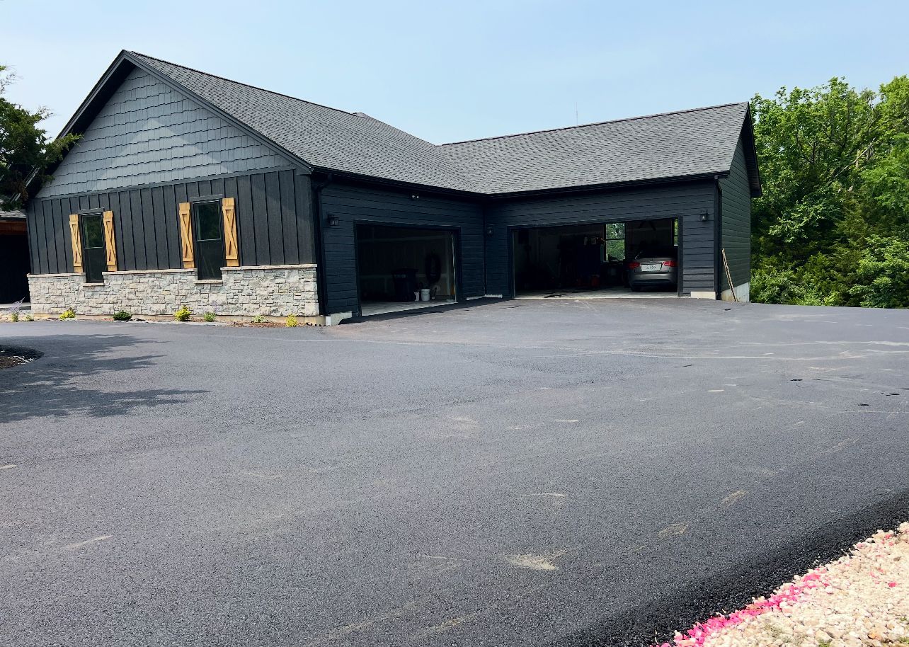 Dark gray barn-style building with open bays and fresh black asphalt driveway under a blue sky