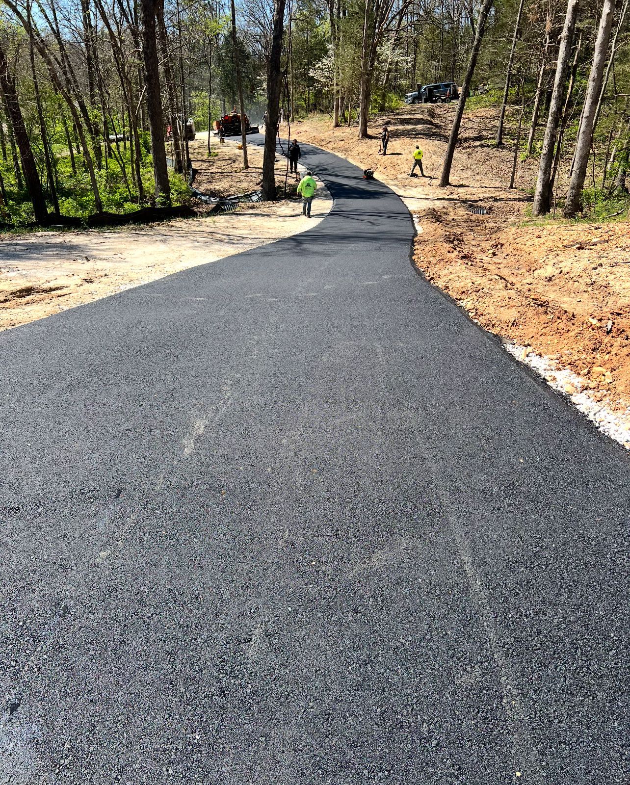Fresh black asphalt path winding through a wooded area with a person and a dog in the distance
