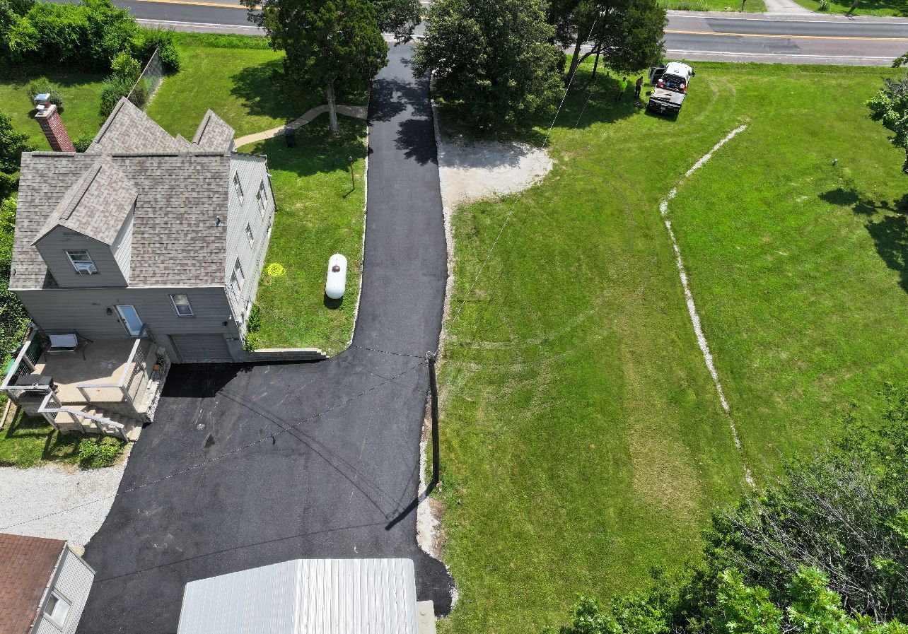 Aerial view of a house, black driveway, parked white car, and green lawn beside a road.