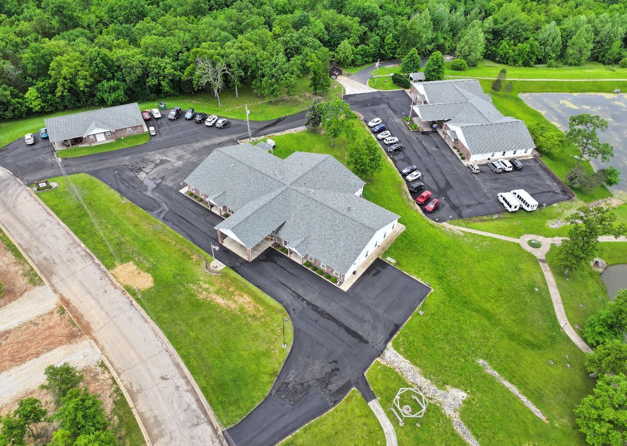 Aerial view of a gray-roofed building with black parking lots beside trees and a road.