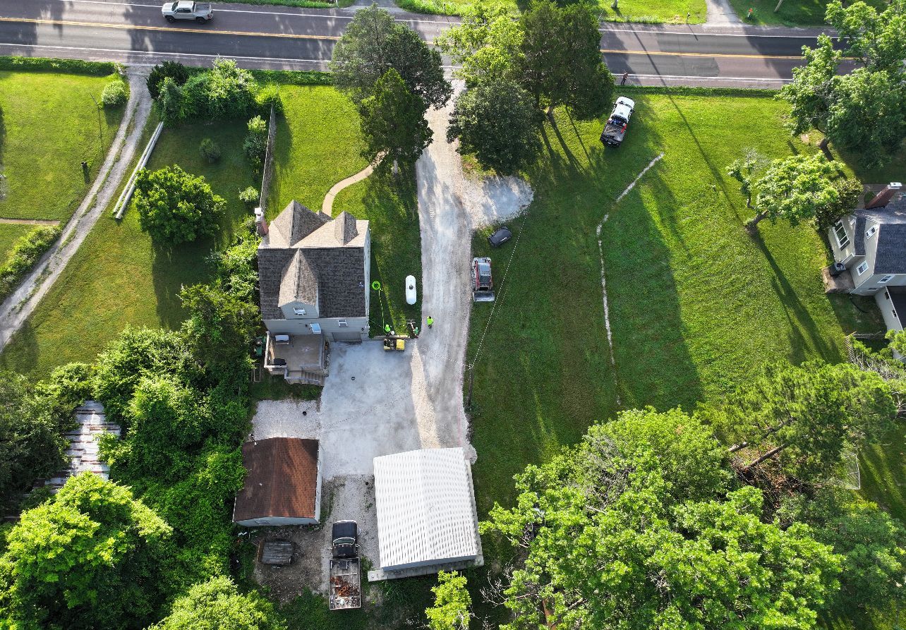 Aerial view of a rural house, dirt driveway, and green fields beside a road