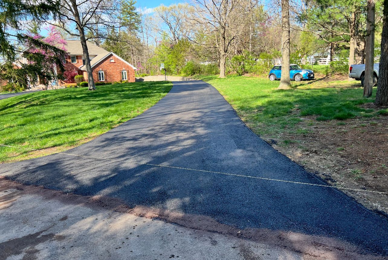 Freshly paved driveway curving through a green yard with a brick house and trees in the background