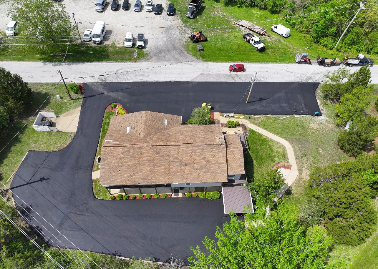 Aerial view of a tan-roofed house with a curved driveway, surrounded by green trees and parked cars.