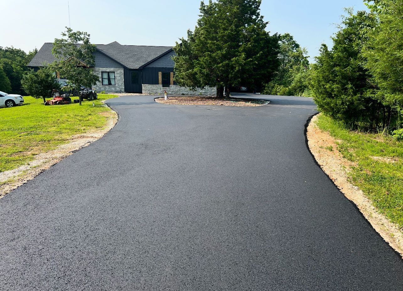 Freshly paved driveway curving toward a gray house with trees and lawn on a sunny day