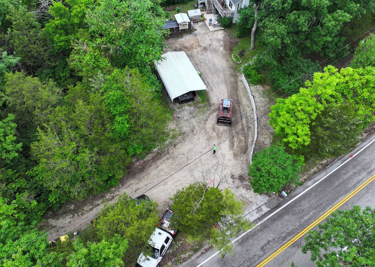 Aerial view of a muddy roadside clearing with parked vehicles, a white canopy tent, and dense green trees.