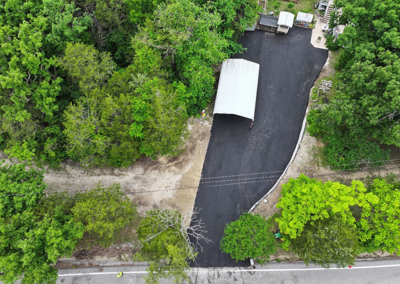 Aerial view of a small white-roofed shed by a dark paved driveway surrounded by dense green trees.