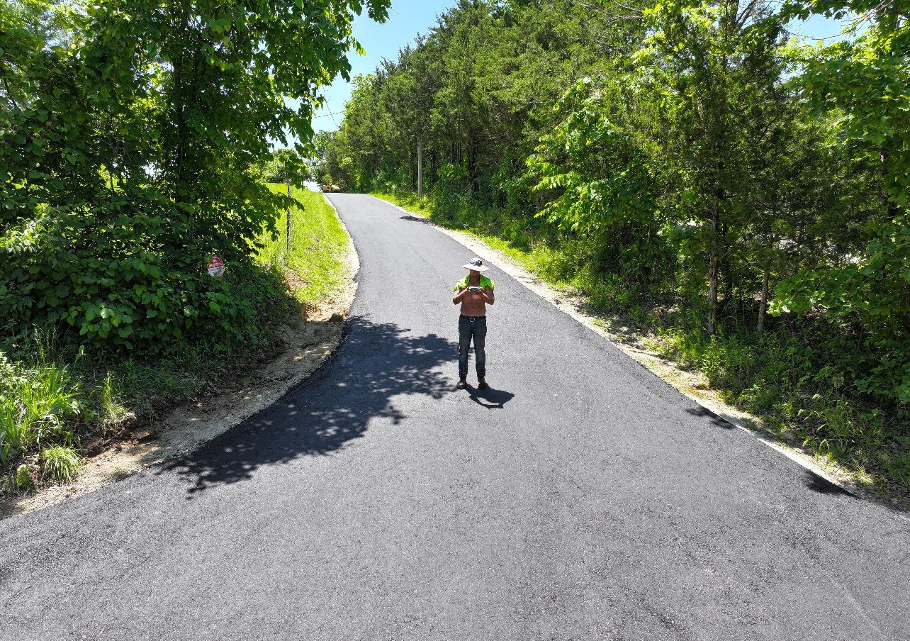 Person standing on a sunlit paved road surrounded by dense green trees