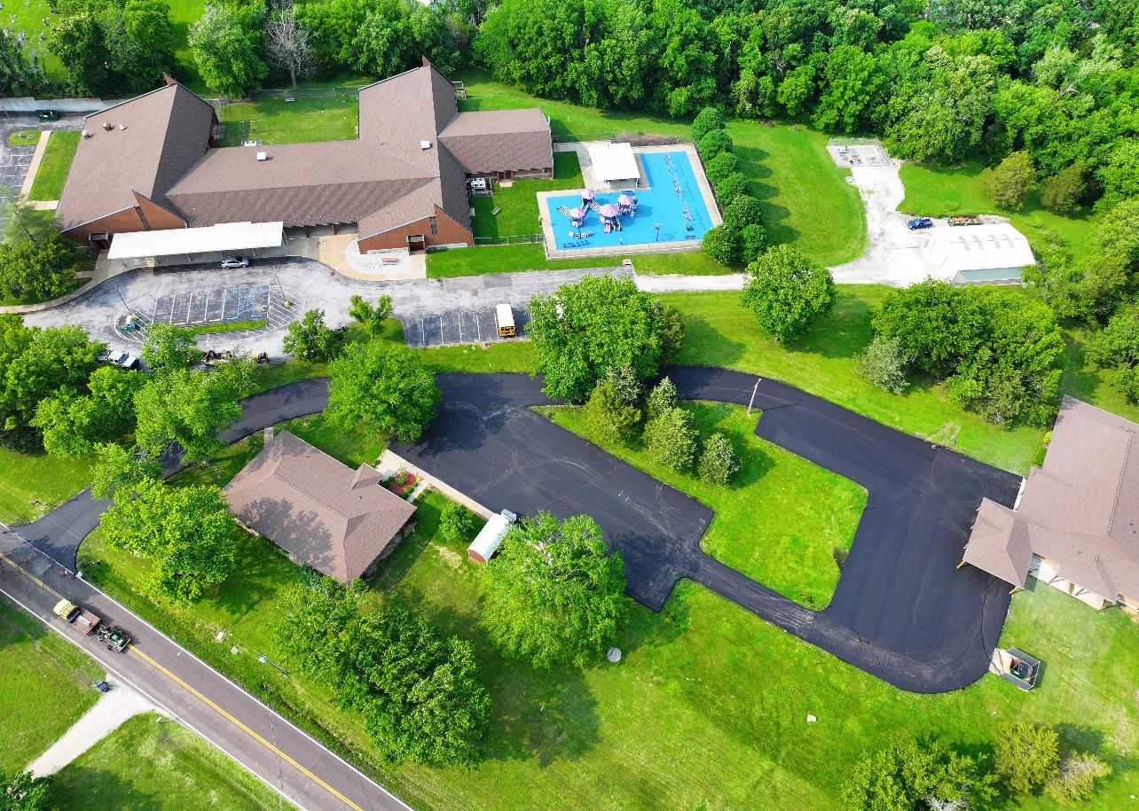 Aerial view of a suburban home with a blue swimming pool, driveway, and surrounding green lawns and trees.