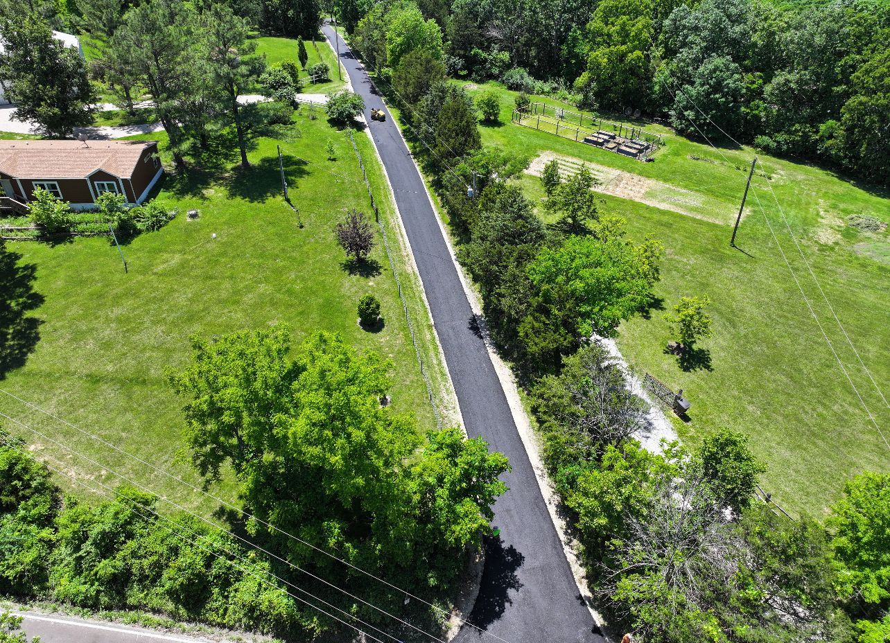 Aerial view of a winding paved road through green fields and trees, with a small house at left.
