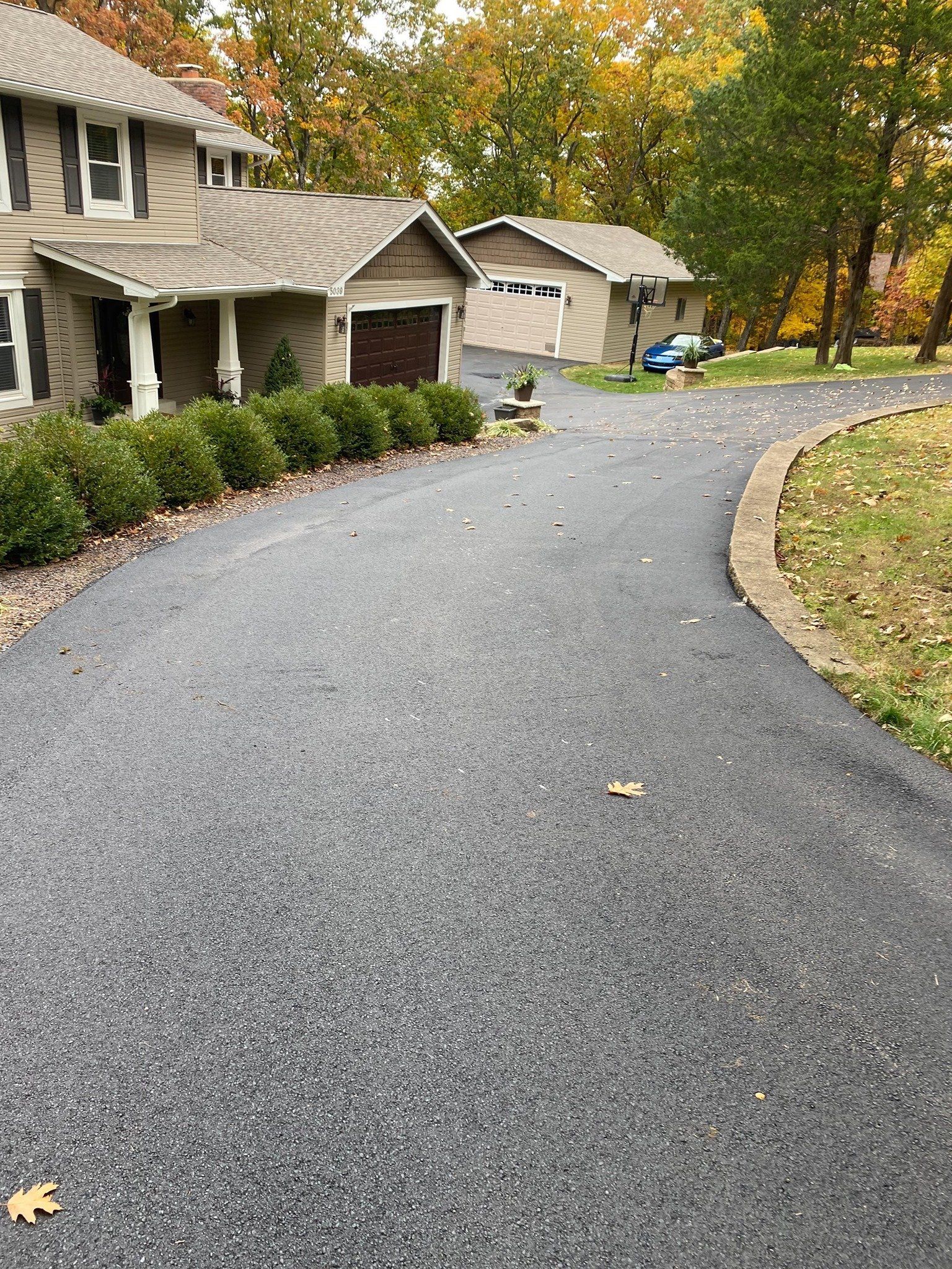Curved driveway beside a house and garage, lined with shrubs and autumn trees on an overcast day